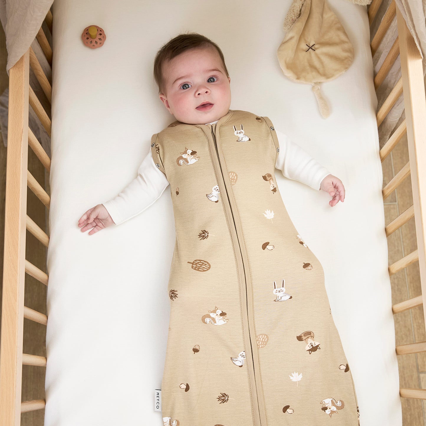 Baby lying in a crib wearing a beige sleep bag with animal patterns.