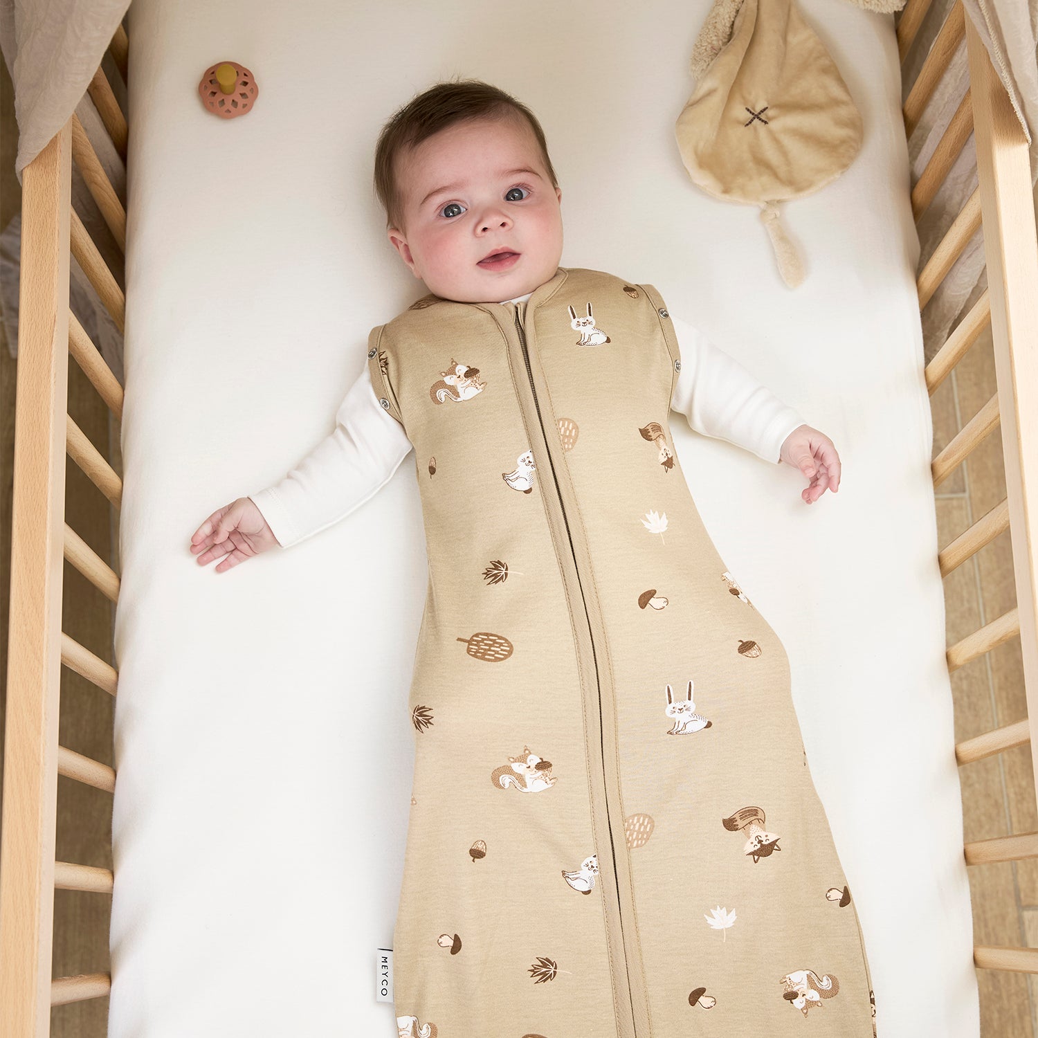 Baby lying in a crib wearing a beige sleep bag with animal patterns.