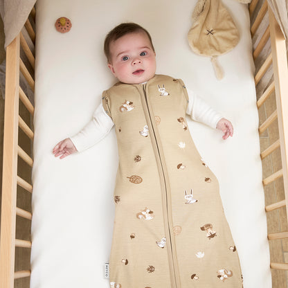 Baby lying in a crib wearing a beige sleep bag with animal patterns.